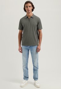 Kortsleevet, olivengrønn polo med glidelås i halsen, kombinert med lyseblå jeans og hvite joggesko. Enkelt design, laget av bomull.