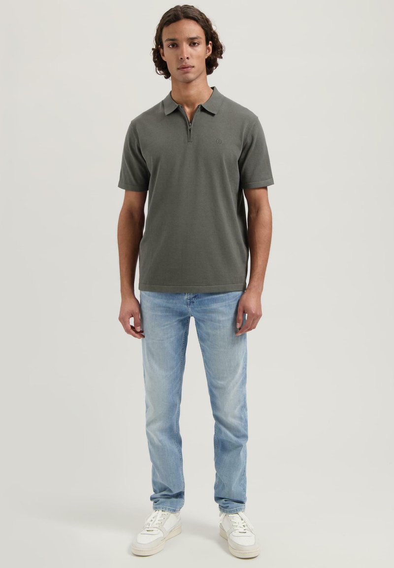 Kortsleevet, olivengrønn polo med glidelås i halsen, kombinert med lyseblå jeans og hvite joggesko. Enkelt design, laget av bomull.
