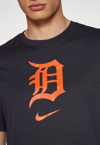 Juodas medvilnės marškinėliai su oranžiniu "D" logotipu ir Nike swoosh. Trumpi rankovės ir apvalios kaklo iškirptės dizainas. Lygus paviršius.