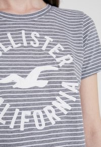 Grå randig t-shirt med vit cirkulär logotyp som står "Hollister California" och en broderad mås-grafik. Tillverkad av bomull, med rundad hals och korta ärmar.
