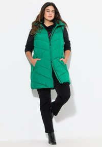 Gilet puffer verde con cappuccio, caratterizzato da un design trapuntato, chiusura a zip e tasche laterali, abbinato a un top nero a maniche lunghe e pantaloni scuri.