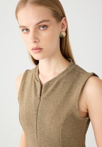 Abito marrone senza maniche con colletto alto e zip frontale, caratterizzato da una texture liscia e un design aderente. La modella indossa orecchini pendenti rotondi.