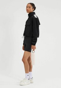 Czarna krótka kurtka z białymi paskami, czarne szorty, białe sneakersy i jasnoróżowa butelka na wodę. Gładki materiał, casualowy sportowy styl.