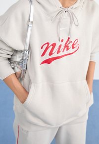 Béžová mikina s kapucí s velkým červen�ým logem Nike, která má šňůrku na kapuci a přední kapsu. Je viditelný stříbrný popruh od tašky.