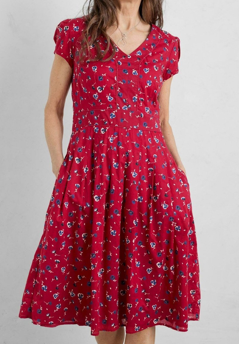 Robe rouge à fleurs avec manches courtes, encolure en V et poches. Présente de petites fleurs bleues et blanches sur une jupe froncée. Texture de tissu douce.