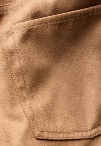 Tissu en suede marron avec une poche avant et des coutures visibles. La texture est douce et légèrement texturée avec une finition mate.