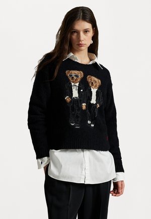 Donna che indossa un maglione nero con due orsetti in abito, sovrapposto a una camicia bianca e pantaloni neri, su uno sfondo bianco uniforme.