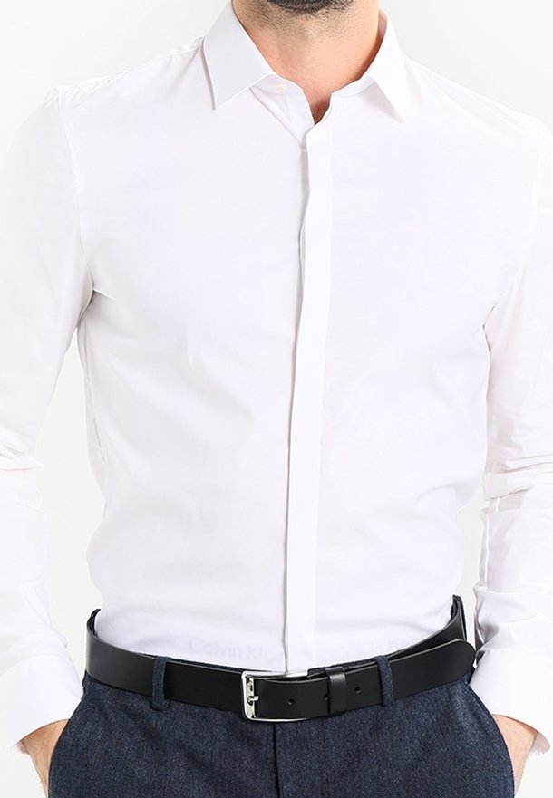 Biała koszula z długim rękawem o dopasowanym kroju, z klasycznym kołnierzykiem i zapinana na guziki z przodu. Zestawiona z czarnym skórzanym paskiem i ciemnymi spodniami.