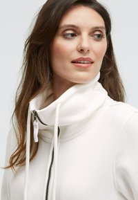 Weißer Zip-Hoodie mit hohem Kragen, ausgestattet mit einem kontrastierenden Reißverschluss und Kordeln. Glatte Textur mit taillierter Passform.