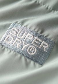 Lys grøn stof med et tekstureret, syet label, der viser "SUPER DRY" i hvid tråd. Inkluderer et cirkulært varemærkesymbol.