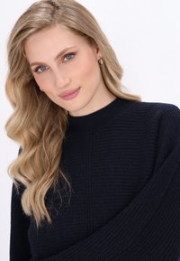 Marineblaues Rippstrickpullover mit hohem Kragen und weiten Ärmeln. Das Model hat lange wellige Haare und trägt kleine silberne Creolen.