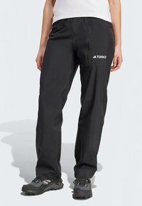 Oseba v črnih prostih hlačah z logotipom "Terrex" na stegnu in sivih pohodniških čevljih, ki stoji pred enobarvno svetlo ozadjem.