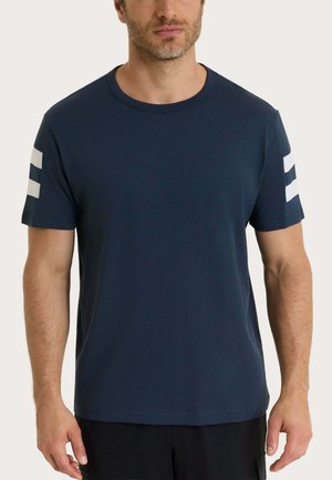 Uomo che indossa una maglietta blu navy a maniche corte con due strisce bianche orizzontali su ogni manica, in piedi davanti a uno sfondo chiaro e uniforme.