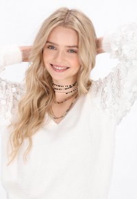 Bijela v-neck pulover s teksturiranim detaljima na rukavima, slojevite višeslojne ogrlice s perlama u raznim bojama, valovita plava kosa.