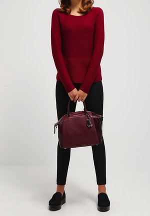 Sac à main en cuir bourgogne avec des accents écailleux et une forme structurée. Il dispose de deux poignées et d'un charme décoratif. Porté avec un pantalon noir.