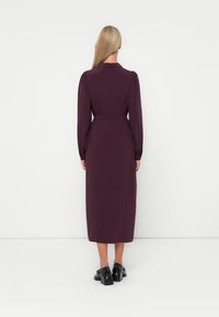 Rochie lungă, de un burgund închis, cu mâneci lungi și talie încinsă, având o textură netedă și un design fluid, purtată cu pantofi negri.