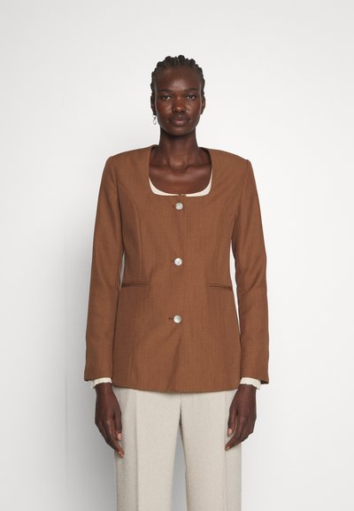 Καφέ κομψό σακάκι με τετράγωνη λαιμόκοψη, ένα μπροστινό κουμπί και πλαϊνές τσέπες, σε συνδυασμό με ανοιχτόχρωμο υφαντό παντελόνι.