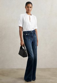Weiße Kurzarmbluse mit Knopfleiste, kombiniert mit hochgeschnittenen, dunkelblauen Jeans im Schlagstil und einer kleinen schwarzen Handtasche.