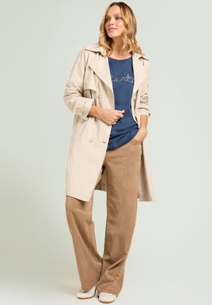 Donna con trench beige, camicia blu e pantaloni marroni a gamba larga, in piedi con una mano in tasca su sfondo chiaro.
