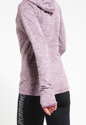 Osoba nosící fialovou mikinu s dlouhým rukávem a černé legíny s vertikálním nápisem "RUNNING", ruka uvolněně podél těla.