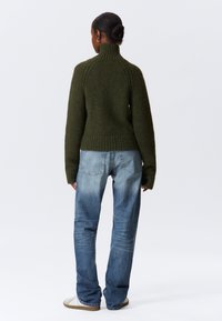Suéter de punto verde oscuro con un cuello acanalado alto, combinado con jeans azules desgastados. Textura suave y corte relajado con mangas largas.
