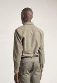 Camisa abotonada en un verde oliva apagado, de material de algodón, con cuello y mangas largas. Combinada con pantalones gris claro y un cinturón trenzado marrón.