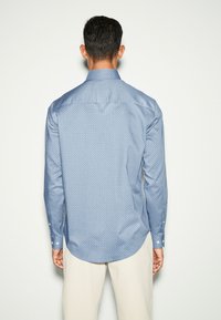 Camicia azzurra a maniche lunghe con un piccolo motivo geometrico, colletto a punta con bottoni, e bottoni bianchi sui polsini. Tessuto dalla consistenza liscia.