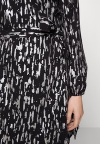 Robe noire avec motif abstrait argenté, manches longues et détail noué à la taille. Tissu lisse et coupe légèrement ample.