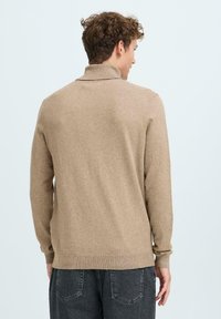 Beige kötött pulóver magas gallérral és hosszú ujjakkal. Bordás mandzsettákkal és szegéllyel rendelkezik. Sötétszürke farmerral viselve, hátulról látható.
