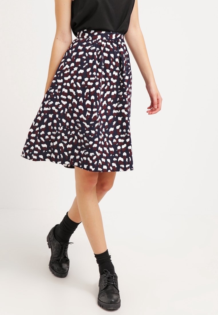 Femme portant une jupe à motifs longueur genou, avec des chaussettes noires et des chaussures noires à lacets, debout devant un fond clair uni.