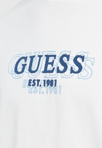 Tela blanca con textura de capas de texto bordado azul "GUESS" y "EST. 1981" en estilos en negrita y contorneados.