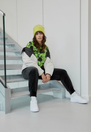 Uldet hvid jakke, neongrøn grafisk t-shirt, sorte velourbukser og hvide sneakers. Model sidder på trapper med glasgelænder.
