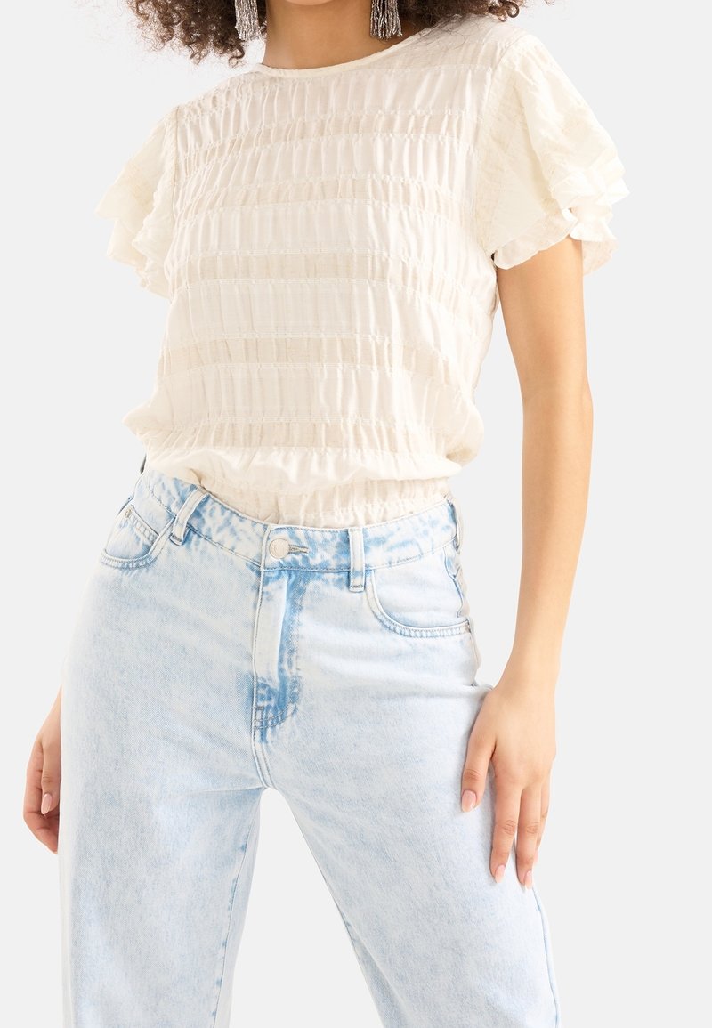 Blouse crème claire à manches courtes avec manches volantées et rayures texturées, associée à un jean en denim bleu clair taille haute avec des taches délavées.