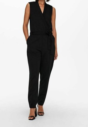 Kobieta ubrana w czarny, bezrękawnikowy kombinezon z zakładanym przodem i przewiązanym w talii, zestawiony z czarnymi sandałami na wysokim obcasie z odkrytymi palcami.