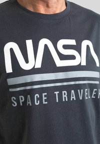 T-shirt in cotone grigio scuro con grande logo bianco "NASA" e strisce argentate di accento, con la scritta "SPACE TRAVELER" sotto. Maniche corte e collo rotondo.