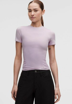 Jeune femme aux cheveux attachés portant un top près du corps violet clair à manches courtes et un pantalon noir taille haute, debout devant un fond uni.