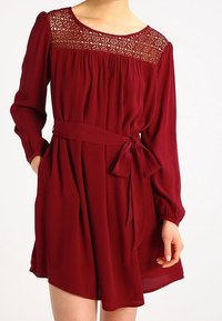 Maroon mekko, jossa on pitkät hihat, pitsiyksityiskohtia olkapäissä, rypistetty vartalo ja solmimisvyö. Pehmeä kangas, polvipituinen malli.