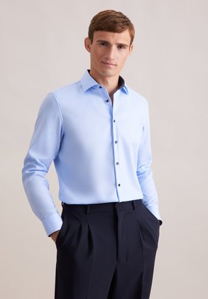 Chemise bleu clair à manches longues avec des boutons noirs et un détail de col contrastant ; associée à un pantalon sombre, en tissu lisse, à coupe ajustée.