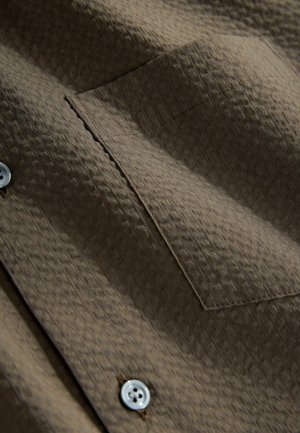 Brun tekstureret skjortestof med en lille brystlomme og hvide knapper, der fremhæver et diagonalt mønster og syningsdetaljer.