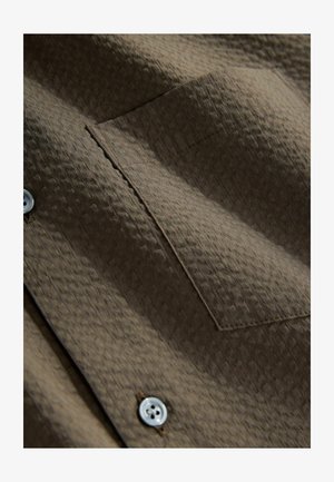 Rjava teksturirana tkanina za srajco z majhnim žepom na prsih in belimi gumbi, ki poudarja diagonalni vzorec in detajle šivanja.