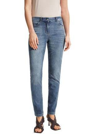 Femme portant un jean slim bleu, un top beige et des sandales à talons noirs avec des brides tressées, debout sur fond blanc.