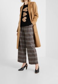 Frau trägt einen Kamelfarbigen Mantel, einen schwarzen Pullover mit drei sandfarbenen Herzen, braune karierte breite Hosen und schwarze mit Nieten besetzte Slingback-Pumps.
