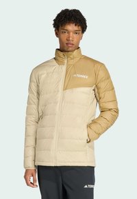 Beige and brown puffer jacket with a high collar, zip closure, and quilted texture. Features branding on the upper left chest.