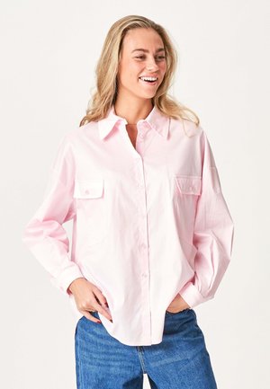 Smilende kvinde med blond hår iført en løs lyserød skjorte med knapper og blå jeans, hænderne i lommerne.