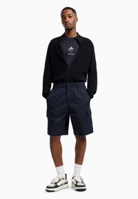 Zwarte ritsvest, marineblauwe cargoshorts, witte sokken en zwart-cremekleurige sneakers. De outfit heeft een casual, ontspannen uitstraling met functionele details.