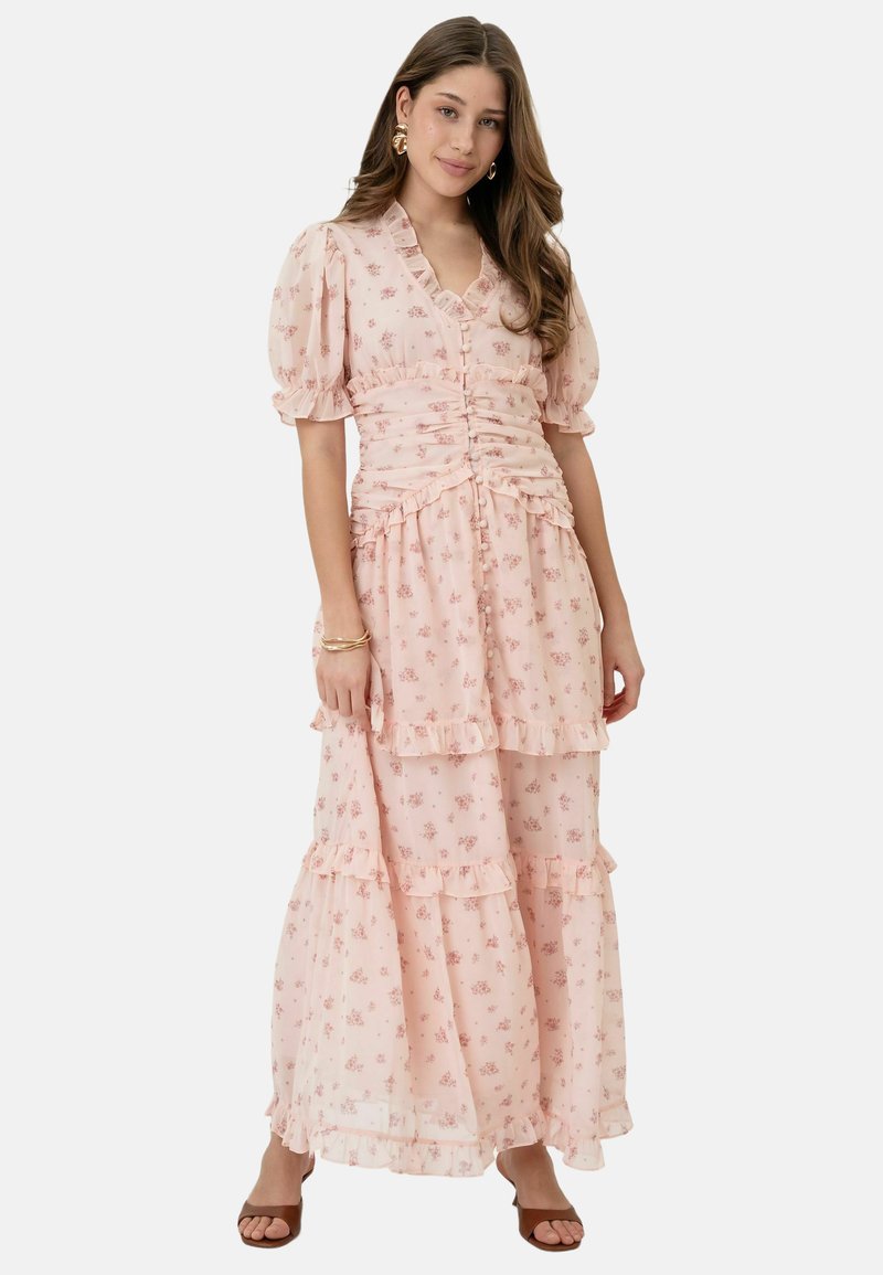 Jeune femme portant une longue robe rose à imprimé floral, manches bouffantes, volants, et des sandales marron ouvertes aux orteils, debout sur fond blanc.