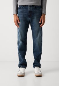 Man wearing dark blue jeans, gray long-sleeve shirt, and white sneakers standing against a plain white background.
