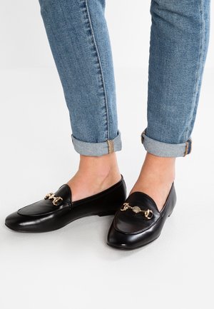 Zwarte leren loafers met een spitse neus, voorzien van een gouden kettingaccent op de wreef. Gekocht met opgerolde denim jeans.