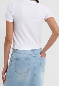Žena v bílém crop topu s krátkým rukávem a světle modré džínsové sukni s kapsami vzadu, pózující s jednou rukou v kapse.