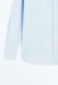 Jasnoniebieska koszula z długim rękawem, wykonana z fakturowanej tkaniny, z zapięciem na guziki z białymi guzikami i zaokrąglonym dołem.